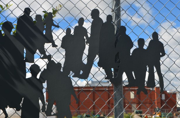metal cutout figures of people walking, alongside a chainlink fence as part of an art installation