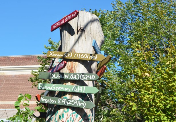 an old tree trunk has been turned into a support for many little arrow shaped signs that point the direction to cities around the world along with how many kilometres it is to those cities. 
