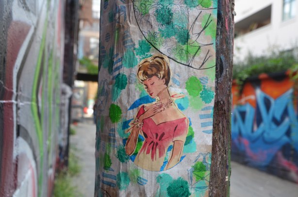 an old fashioned type drawing in colour of a woman with short brown hair and a red v neck dress, from the waist up, eyes closed, face looking slightly downwards, on paper on a pole in a lane. 