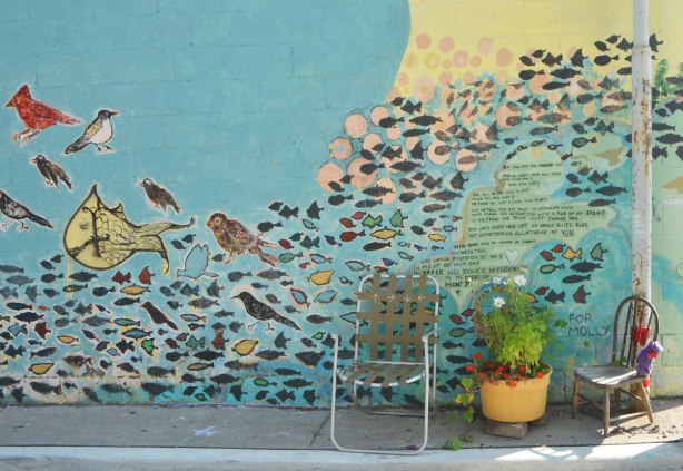mural on a wall with lots of birds and fish and a poem as a memorial to a cat, with two chairs in front of it as well as a yellow planter with flowers in it. 
