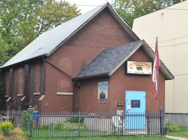 brown brick builgin, one storey with peak roof, small veranda in front, blur front door, Canadian flag out front, sign above door says Royal Antidiluvian Order of Buffaloes. 