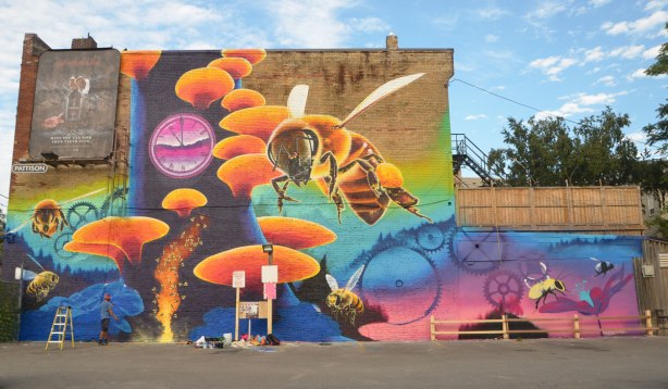 large mural, Riverside Pollinator mural, by Nick Sweetman, of a large bee, a clock, clock gears, a few small bees, a honeycomb and a flower and a very large tree