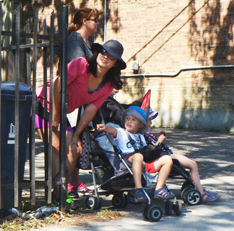 a woman in a pink top and a black wide brimmed hat leans down to talk to her son who is sitting in a stroller. He's pointing down the sidewalk. They are watching a parade go by. 