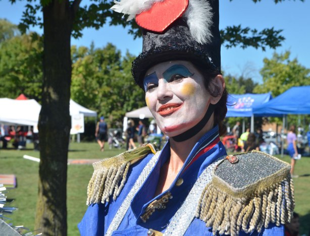 a woman white painted white face, yellow cheeks and red lips, wearing a costume in blue with padded shoulders and a fringe around the shoulders, top hat with but white feather and a big red heart 