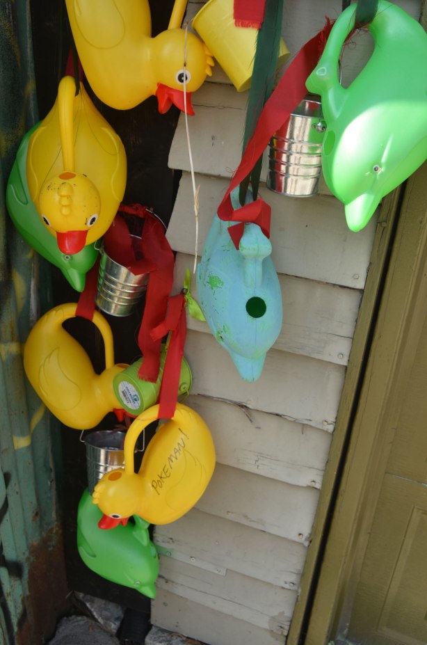 watering cans hanging along the side of a garage in an alley, many of them are in the shape of yellow rubber duckies and one is a light blue fish shape. A few are little silver coloured metal buckets. 
