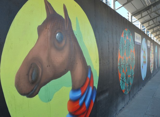 large circle on black wooded construction hoardings.  In the circle, painted light green is a brown horse head and neck, with a red and blue striped scarf around its neck, painted by birdo 