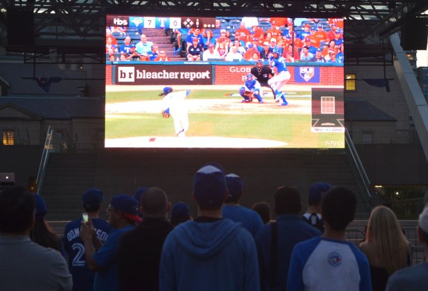 people watching the Blue Jays baseball team playing a game on a large TV screen outside.