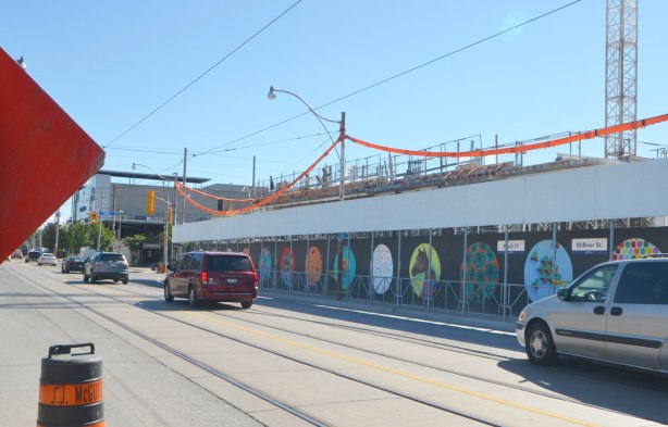 A street, Dundas East, with construction hoardings painted with street art pictures inside large circles ona black background, cranes in the background. A couple of cars