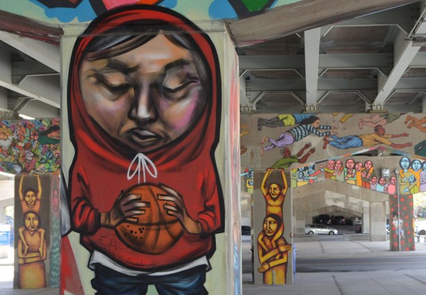 elicser painting of a basketball player in a red hoodie on a pillar in underpass park, other pillars and bents are in the background, all of which have been painted with pictures of people, painted by different street artists. 