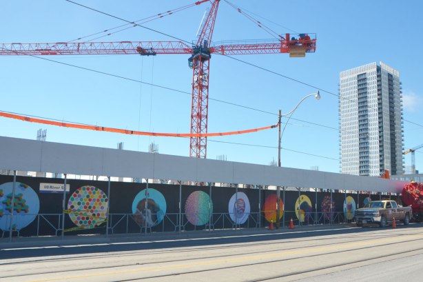 A street, Dundas East, with construction hoardings painted with street art pictures inside large circles ona black background, cranes in the background. 