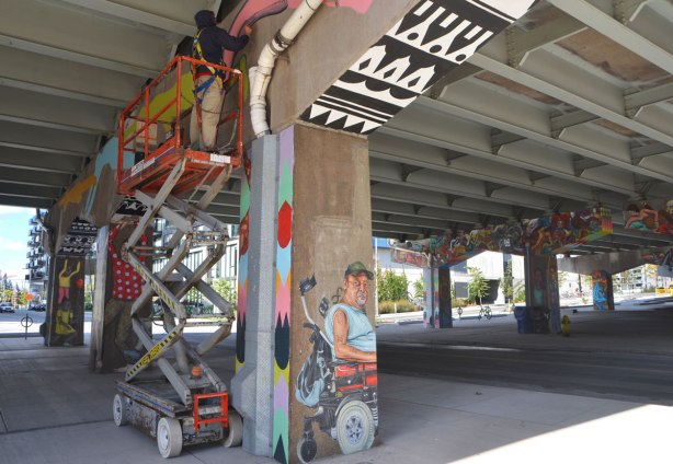 a street artist (Troy Lovegates) is on a lift and painting a mural on the concrete supports on an underpass. Other supports are in the picture, they have already been painted. 