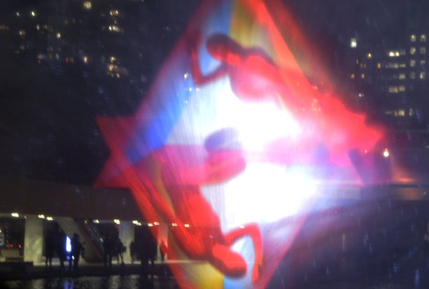 images of people in triangular shapes projected onto a wall of water as part of an art installation, night time, lights of buildings in the background.