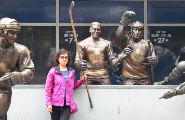 a woman in a pink jacket stands in front of a statue of a hockey player, Tim Horton. She is holding onto his hockey stick. There are a couple of other statues in the picture too.