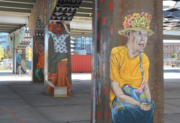 street art murals on 4 bents holding up the ramp of a highway, underpass park, the paintings are of people, in the foreground is a woman with a yellow T-shirt and a hat with little flowers