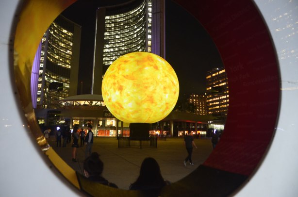 looking through the O of the Toronto sign to the yellow and orange projections on the large globe in front of City Hall, art installation from Nuit Blanch