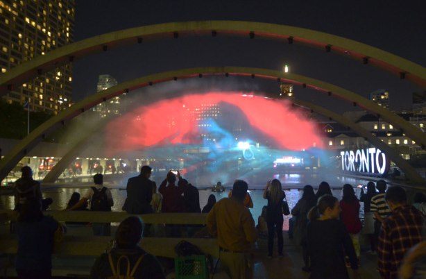 people standing around with their backs to the camera in the foreground, with a picture projected onto a wall of water under the arches at Nathan Phillips square in the background. The picture is of a blue woman on a red background.