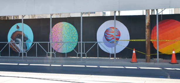 construction hoardings, painted black and on that, large circles with paintings on them.  A bird head, one with green and pink squiggles, and one with a zebra striped sphere in orange and blue and the fourth with diagonal stripes making a gradient from yellow to dark red