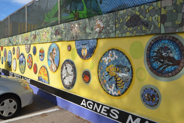 A wall with a chainlink fence above it, a large green construction crane is behind the wire fence. The wall has been covered with a mural, yellow background and mosaic pictures on it. A quote runs along the bottom at ground level, white letters on black background, quote by Agnes MacPhail