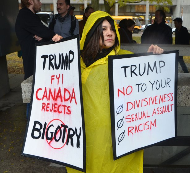 people at a rally protesting against Donald Trump as President of the USA, a woman in a yellow raincoat holds two signs, one says Trump FYI Canada rejects bigotry and the other says Trump no to you divisiveness, sexual assault and racism