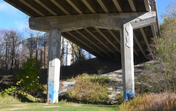 in a park under a bridge, grassy area, some trees on either side, 2 concrete support pillars with street art on the bottom of each. 