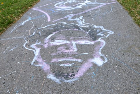 large chalk drawing of a man's face in black, white and pale purple, on a paved path in a park 