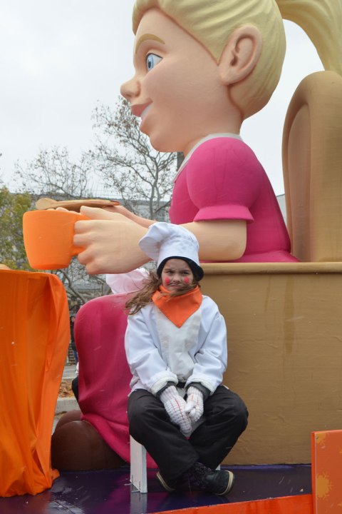 Santa Claus parade - a girl is looking cold, sitting on a float in chef's jacket and hat and large orange scarf. She is sitting beside a large sculpture of a woman sitting on a wood chair with an orange cup in her hand 