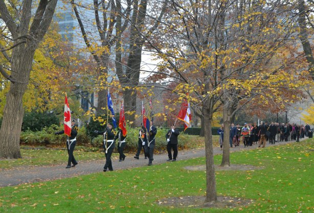 colour party of 6 men carrying flags lead a procession through the park in front of the parliament buildings at Queens Park, autumn and most of the leaves are off the trees.
