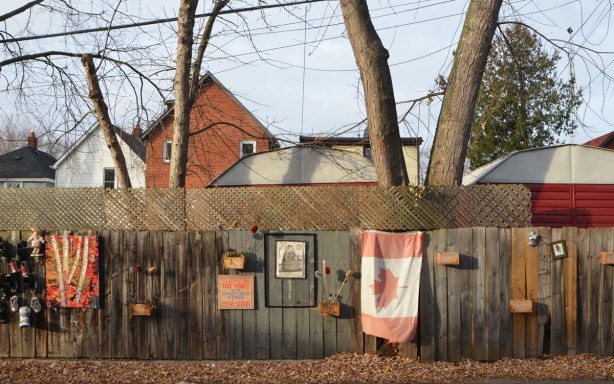 decorated wooden fence on Craven Road, a road with houses only on one side of the street, fence is decorated with a faded Canadian flag, a picture of Queen Elizabeth I, some wood planters, a painting of birch trees in autumn, old shoes, and a sign that says Craven road FEnce, 100 years, 1916 to 2016 