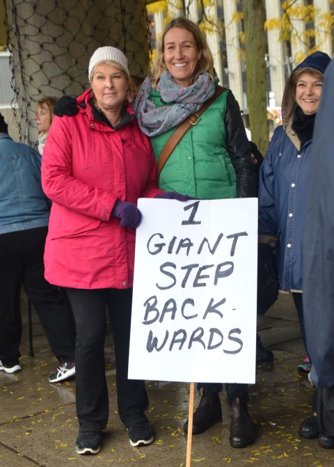 three women at a protest rally against Trump and US politics, holding a sign that says 1 giant step backwards. Nathan Phillips Square, Toronto, rainy day