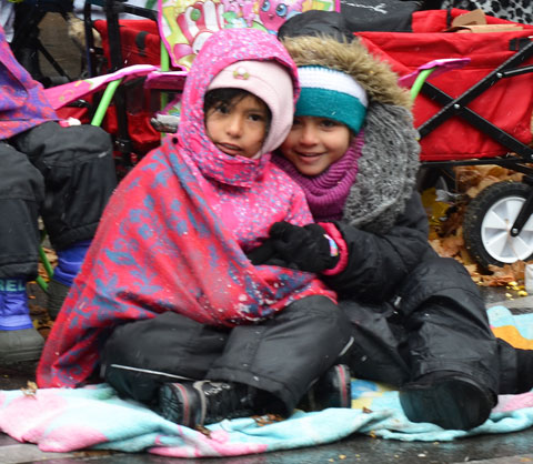 Santa Claus parade - two girls huddle under blankets and parkas while sitting on the sidewalk waiting for the parade to start 