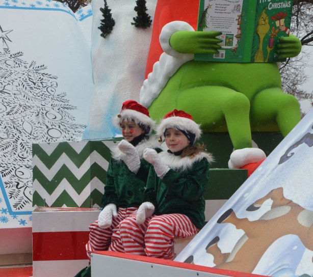 two kids dresses as Christmas elves in green, red and white wave to passersby as they sit on a float with a large Grinch reading a book, Santa Claus parade 