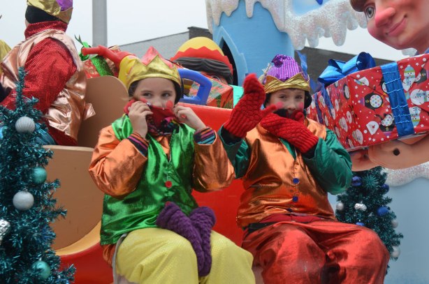 Santa Claus parade - two kids in shiny orange, green, red and yellow costumes, sitting on a float in the parade, they have their hands in mitts covering their mouths and chins trying to keep warm. 