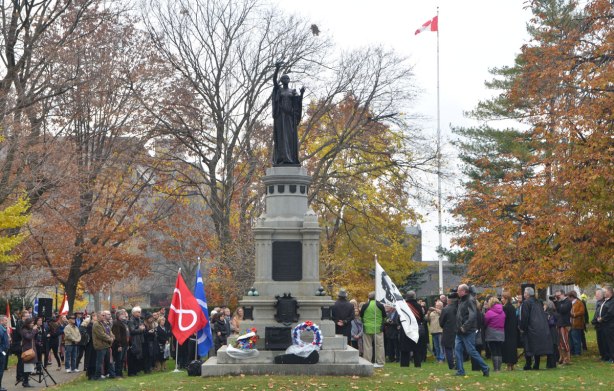 statue, memorial to those who died in the Northwest Rebellion of 1885, to the soldiers on the winning side, at Queens Park, with people around it, a flag with picture of Louis Riel is planted beside the monument,