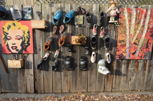 a wooden fence with many things hanging on it - an Andy Warhol Marilyn Munroe print, a painting of autumn birch trees, and many old shoes
