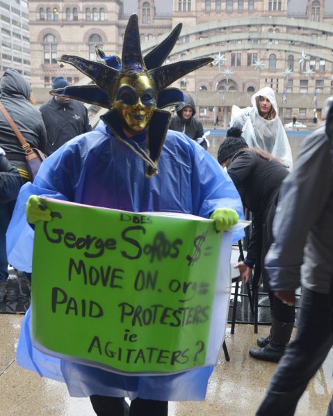 people at a rally protesting against Donald Trump as President of the USA, a person in a mask like a mardi gras mask, and holding a green sign that says Georege Soros move on