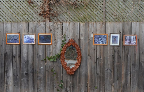 An oval mirror with an ornate wood frame is mounted on an outdoor fence, wood, three small framed pictures hang on both sides of it. 