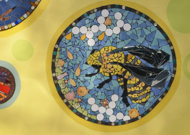 a circular mosaic picture of a bee amongst white and orange flowers on a blue background, all on a yellow wall. Part of a larger mural