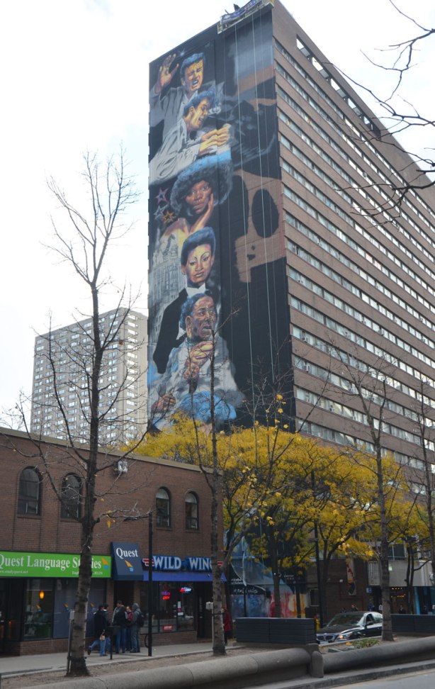 a vertical mural painted on the side of a multi storey building, the mural is a series of faces in a totem pole like arrangement
