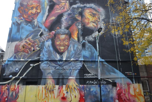 musicians and singers painted in a mural, a black man in a suit, a black man playing a guitar, a man with longish hair singing into a mic with his eyes closed. 