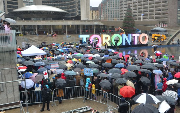 people at a rally protesting against Donald Trump as President of the USA, a wet rainy day, looking out over Nathan Phillips Square with city hall, the 3D Toronto sign and many umbrellas