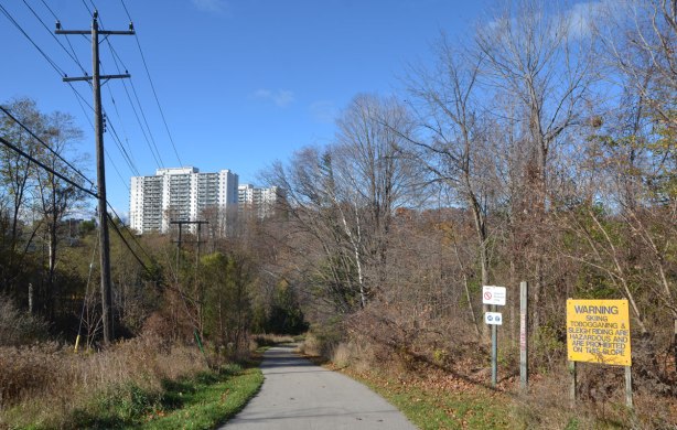 paved path leading down a hill to a ravine park, apartment buildings in the distance. Sign beside path says no skiing or sleigh riding on the slope.