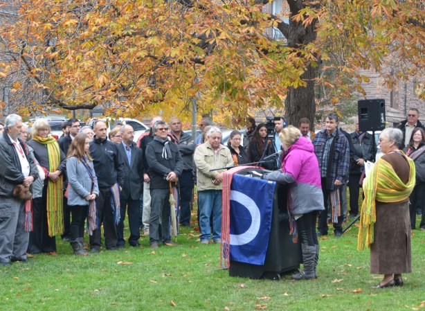 a woman in a pink jacket stands behind a podium draped with the Metis flag, speaking to a group of people outdoors at QUeens Park on Louis Riel day.