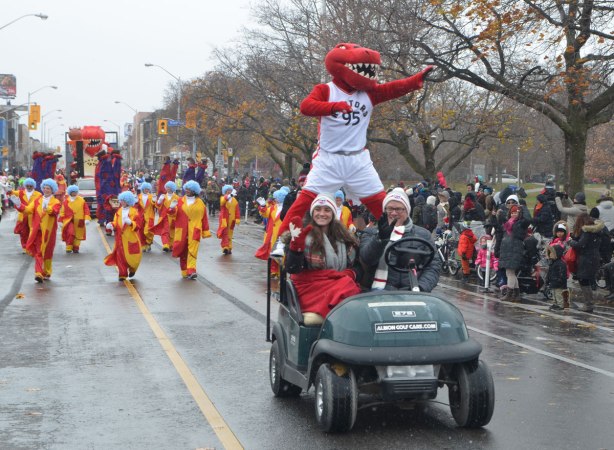 Santa Claus parade - the mascot for the Toronto Raptors basketball team is standing on the back of a golf cart driven by two people, others in costume walk behind, people standing on the sidewalks watching the parade go by 