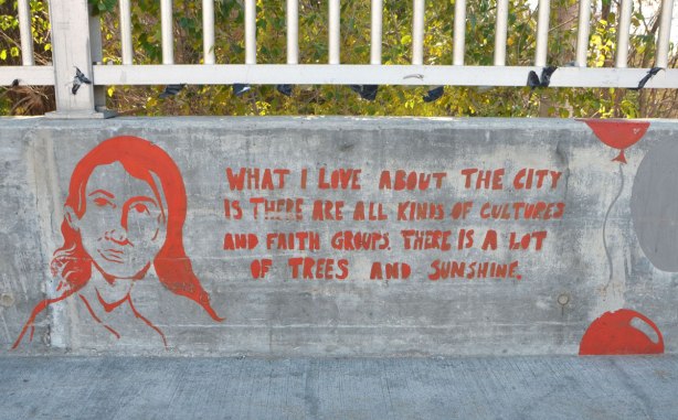 red painting on concrete wall barrier beside sidewalk on bridge over Highland Creek, in red, a girl's head and the words, What I love about the city is there are all kinds of cultures and faith groups. There is a lot of trees and sunshine.