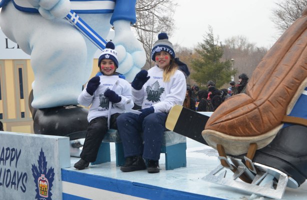 Santa Claus parade - two kids wearing Toronto Maple Leafs sweatshirts and toques sitting on a float with a loarge replica of Carlton the Leafs mascot. The skate and end of the stick of a large statue of a hockey player is also in the picture. 
