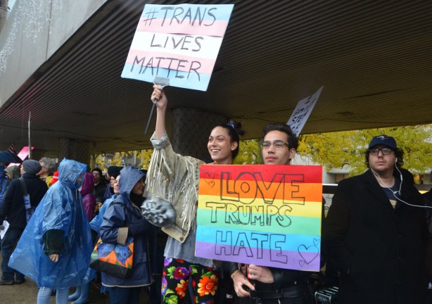 people at a rally protesting against Donald Trump as President of the USA, a young couple with placars. One says Trans lives matter and the other is painted in rainbow stripes with the words Jove Trumps Hate written on it.