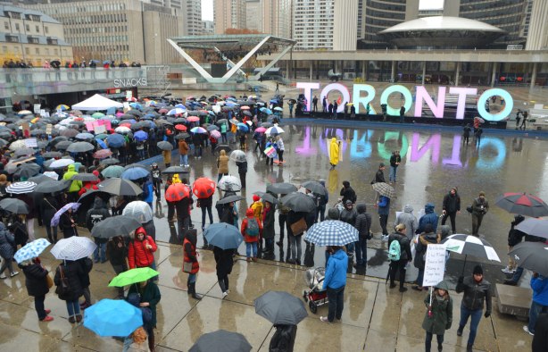 people at a rally protesting against Donald Trump as President of the USA, a wet rainy day, looking out over Nathan Phillips Square with city hall, the 3D Toronto sign and many umbrellas