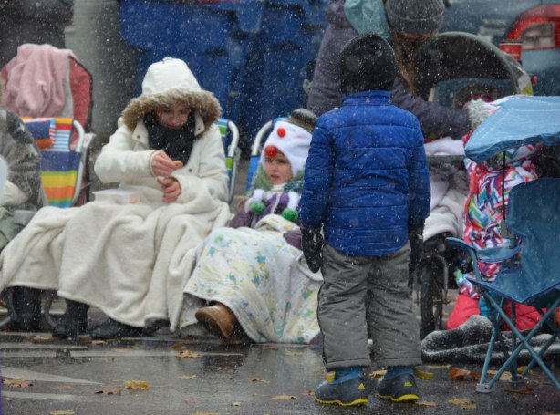 Santa Claus parade - a young boy stands with his back to the camera. He is in front of people sitting on the sidewalk waiting for the parade to start. It is snowing and people are all bundled up. In this case, a mother and her daughter are dressed in white jackets and have a white blanket over their knees. 