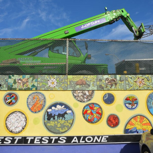 A wall with a chainlink fence above it, a large green construction crane is behind the wire fence. The wall has been covered with a mural, yellow background and mosaic pictures on it. A quote runs along the bottom at ground level, white letters on black background,