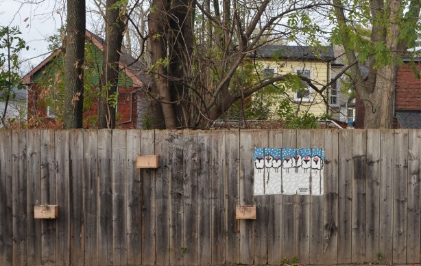 a painting of a line of white sheep on a snowy field, on a fence, outdoors, with houses and backyards beyond the fence, some trees too but they have lost their leaves because it is late autumn